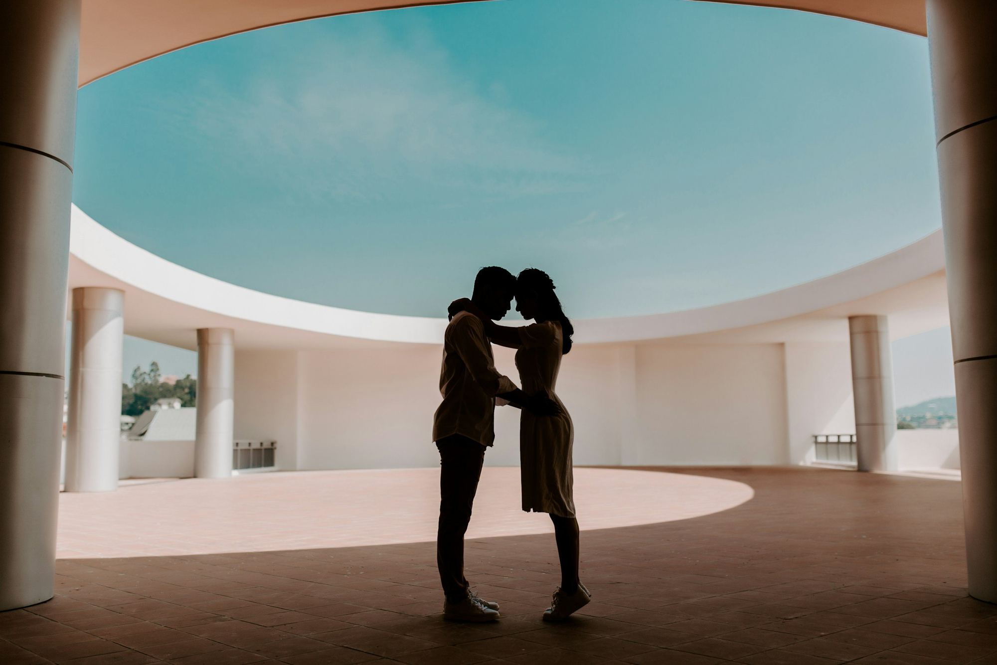 Silhouette of a couple embracing beneath an open circular roof against a blue sky
