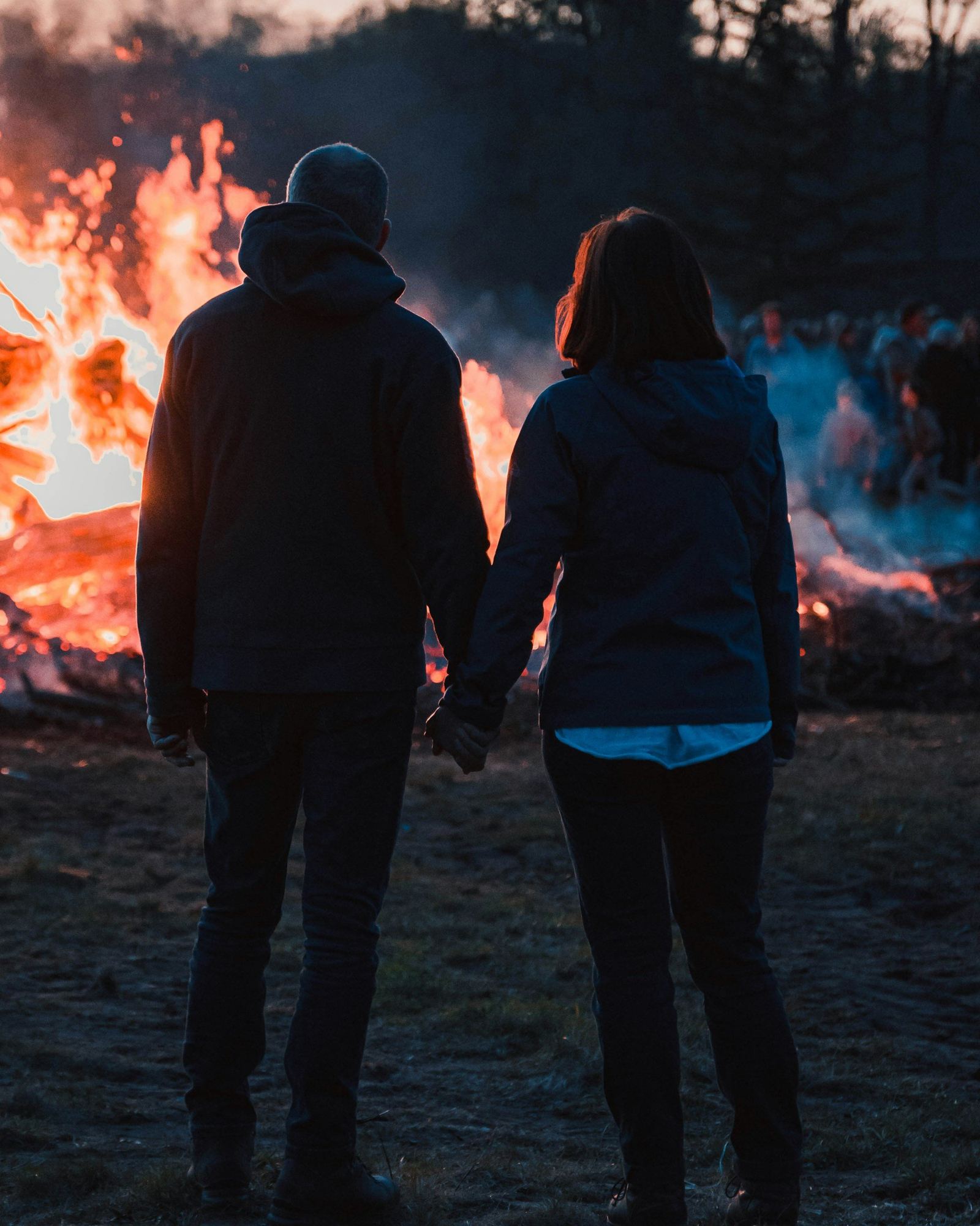 A couple holding hands, seen from behind, in front of a bonfire at dusk